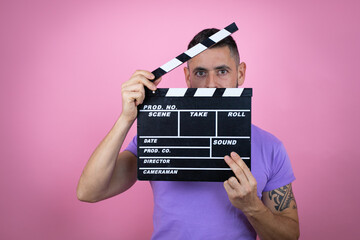 Young handsome man wearing casual t-shirt over pink background holding clapperboard very happy...