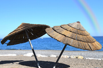2 parasols en paille sur la plage avec un arc en ciel au-dessus de la mer