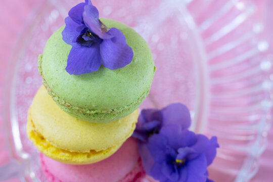 Three colorful french macarons decorated with fragile violet flowers on transparent glass plate.
