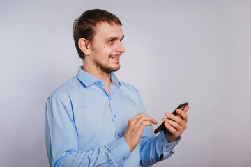 A guy in a blue shirt holds a smartphone on a white background. A male office worker is typing text on the phone. Photo in studio