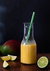 A fresh mango and lime smoothie in a glass milk bottle shape jar, with lime segments and mango fruits on table surface. Copy space available with dark background and wood table.