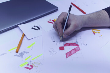 Graphic designer designs a logo against a background of sketches and drawings on a table. Printed logos on paper in a studio with a laptop.