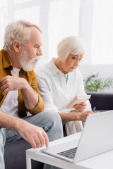Senior woman with calculator and pen sitting near husband and laptop on blurred foreground