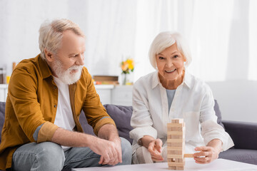 Smiling senior woman playing blocks wood tower game near husband on couch