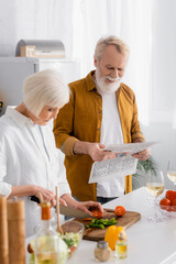 Elderly man reading newspaper while wife cutting vegetables near wine in kitchen