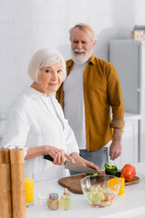 Senior woman looking at camera while cutting paprika near near husband on blurred background