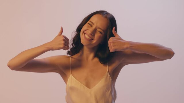 happy young woman in silk camisole dress showing thumb up isolated on grey