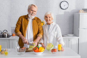 Smiling senior woman standing near husband cutting cabbage and orange juice in kitchen