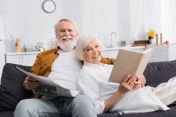 Cheerful elderly couple reading holding book and newspaper on couch