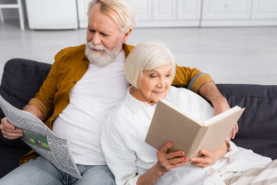 Smiling elderly couple reading book and news on couch at home
