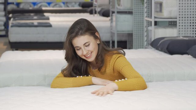 Beautiful happy woman examining orthopedic mattress on sale. Cheerful female customer choosing new bed and mattress to buy at furnishings store. Bedding concept