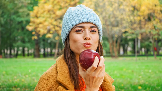 joyful woman looking at camera while eating juicy apple in park