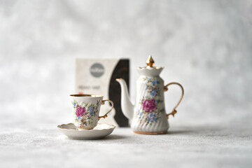  A porcelain cup and teapot next to a package of herbal tea on a gray textured background. Selective focus. Copy space.