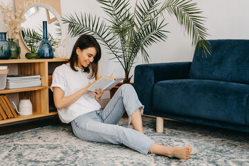 Happy young woman reading a book at home, sitting on the floor in living room.
