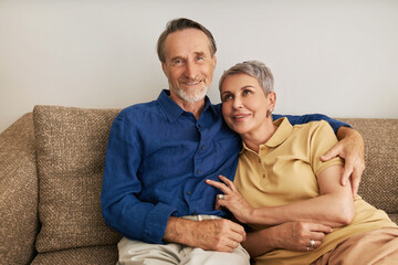 Portrait of smiling senior couple sitting on a couch in a living room. Two positive mature people embracing at home.