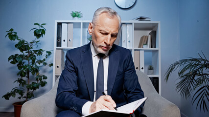 Senior businessman in suit writing on notebook in armchair