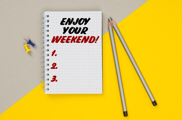 On a yellow-gray background are pencils, buttons and a notebook in which it is written - ENJOY YOUR WEEKEND