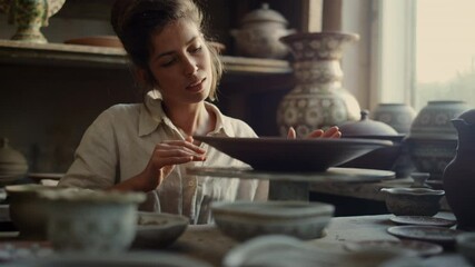 Smiling lady touching plate in pottery. Artist enjoying clay product in workshop