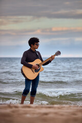 young caucasian teenage boy playing acoustic guitar on the beach. sunglasses and dark clothing. wetting her pants with a wave
