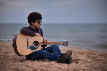 young caucasian teenage boy playing acoustic guitar on the beach. sunglasses and dark clothing.