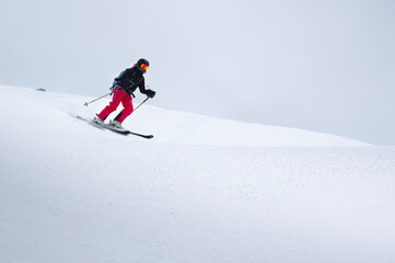 A freerider skier in red pants with a rockzack rides down a snowy slope in cloudy weather. Womens freeride sports background