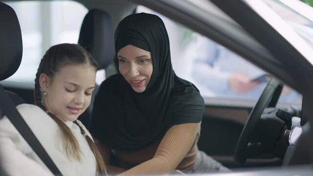 Positive smiling mother in hijab fastening seat belt for daughter sitting in car. Portrait of happy Muslim beautiful woman choosing new automobile in dealership talking with daughter. Slow motion.