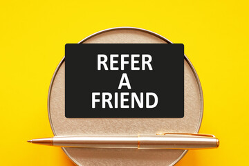REFER A FRIEND - words on a black sheet on a yellow background with round metal stand and metal writing pen. Business, finance and education concept