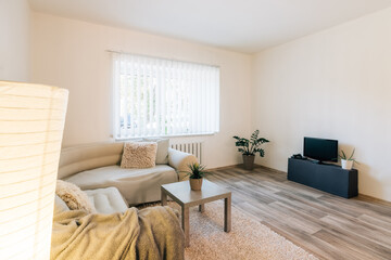 Simply furnished living room in minimalist style.Room has large corner sofa with blankets and pillows, coffee table with plant,lamp,TV table and carpet.There is also large window with interior blinds.