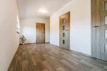 Hallway with various contemporary room door designs, wooden floor and one window. All doors have a wooden texture, glass panels of various shapes and fittings made of matt stainless steel. 