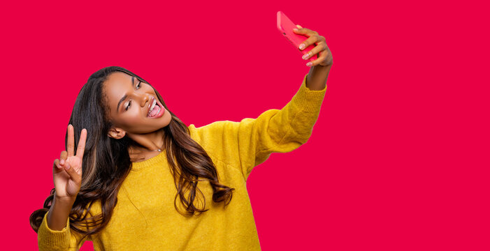 Hello. Portrait of cool cheerful African American girl in yellow sweater having video-call or taking selfie, holding smart phone in hand shooting selfie on front camera isolated on pink background.  