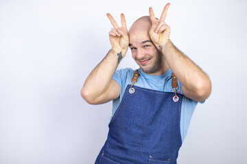 Young bald man wearing apron uniform over isolated white background Posing funny and crazy with fingers on head as bunny ears, smiling cheerful