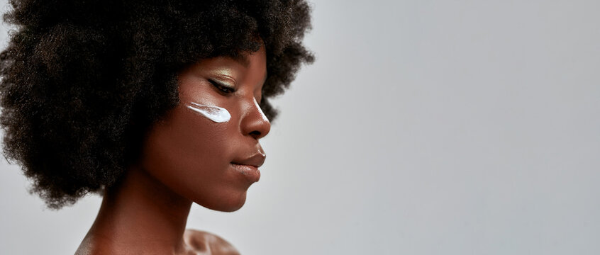 Cropped portrait of beautiful young half naked african american woman with afro hair looking down while posing with cream applied on her face isolated over gray background