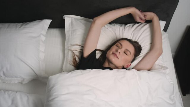  Portrait of relaxed smiling woman wakes up on a white pillow in bed. Portrait of young Caucasian woman resting in bedroom at home. Leisure and relaxation.