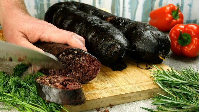 Home kitchen. Chef poorly cuts fresh appetizing blood pudding with blunt kitchen knife on wooden cutting board. Filling spilled out. Vegetables and herbs lies on table. Close-up.