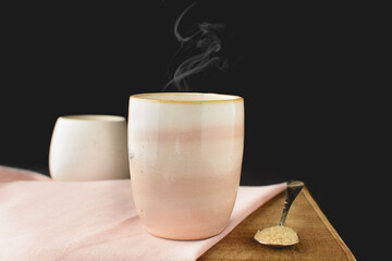 White and pink ceramic cup, giving off smoke, with black background, on a wooden table with light towel and a spoon with sugar beside it