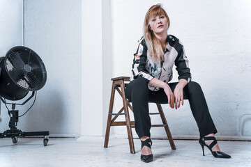 Blonde young woman with defiant eyes and tousled hair on a low wooden stool. Concept: style,...