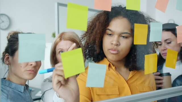 Group of women are talking and writing on colorful sticky notes working with glassboard in office together. Brainstorming and creative work concept.