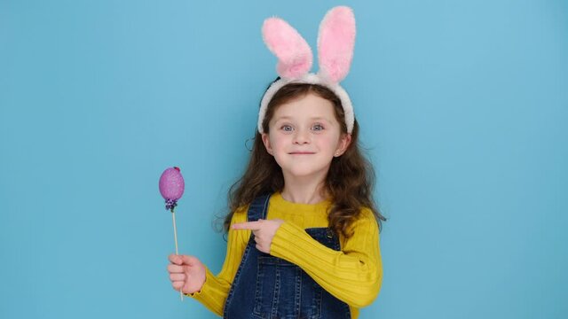Positive cute little kid girl wears pink hare ears and jumper, points small colored eggs grow, makes holiday decorations, models over blue studio background. Look at this decorated Easter eggs! 
