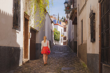 A young blonde female tourist exploring the narrow cobblestone backstreet of the old town (Albaicin or Arab Quarter) in Granada, Andalusia, Spain.