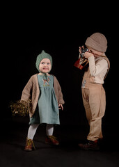 A little boy photographer take a picture with a girl using an old film camera. Children are dressed in cozy retro clothes. Studio isolated on a black background.
