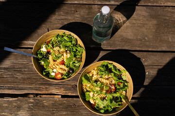 Greek mediterranean salad  with feta cheese, pasta, olives, cherry tomatoes and arugula on a bamboo bowl on a wooden picnic table outdoors