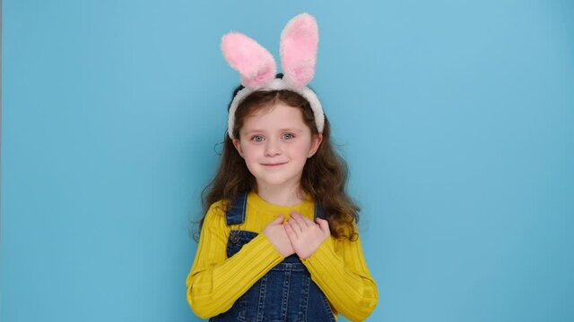 Portrait of smiling cute preschool girl kid in pink bunny fluffy ears put folded palms on heart, isolated on blue studio background. Concept easter and sign of sincere kind person, feeling thankful