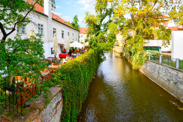 Fototapeta na wymiar A waterfront cafe with outdoor patio along one of the lush garden canals in the Kampa Island area of Prague, Czechia.