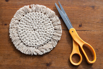 A crochet, or macrame, coaster for a mug on a dark brown wooden desk next to a pair of scissors laying down flat with bright orange handles, February 2021.