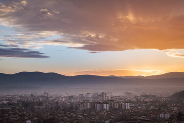 Sun beams shining through the colorful sunset colored clouds over golden lighten city covered by mist