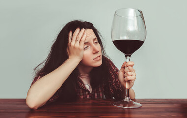 A young caucasian girl in a plaid shirt with tousled hair sits at a wooden table and holds a large glass of red wine.Concept of alcohol abuse, headache, alcoholism, hangover, loneliness and depression