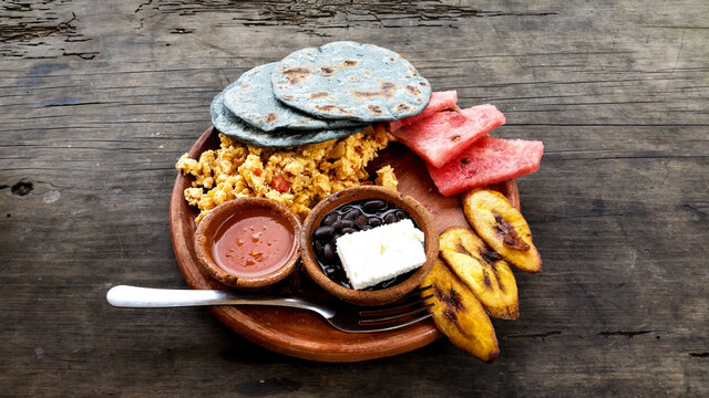 Desayuno tipico Guatemalteco, con frijoles negros con queso, huevos revueltos, platanos fritos, salsa y tortillas negras. Acompa&ntilde;ado de fruta.