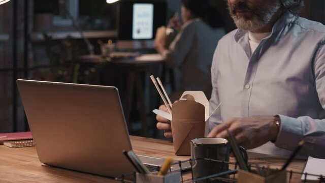 Tilting slow-motion close up of concentrated man looking at laptop screen and eating chinese food from carton box, staying late in office, his colleague working in background
