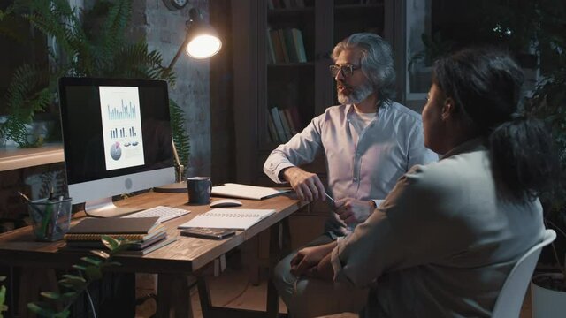 Medium shot of cheerful multi-ethnic coworkers sitting at table in loft office at night, looking at computer monitor and discussing business charts and diagrams