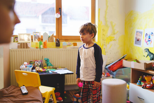 Portrait of small caucasian boy four years old child standing at home in the mess in room wearing pajamas playing and looking to the side surrounded with toys - childhood growing up family concept
